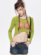 Young woman in a green long-sleeve crop top with a pink butterfly design, paired with a denim mini skirt. Fashionable casual outfit, trendy style.