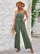 Woman in stylish green strapless jumpsuit, wearing a straw hat and holding a woven clutch, standing against a white stone wall with greenery.