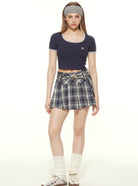 Young woman in a navy crop top and plaid skirt, wearing a headband and leg warmers, showcasing trendy casual fashion.