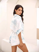 Woman wearing a white oversized shirt with blue abstract patterns, standing in a minimalist setting. Fashionable summer outfit, casual style.