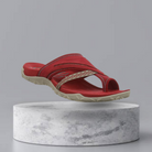 Red orthopedic sandal with arch support, featuring a crisscross strap design and cushioned sole, displayed on a marble pedestal. Perfect for comfort and style.