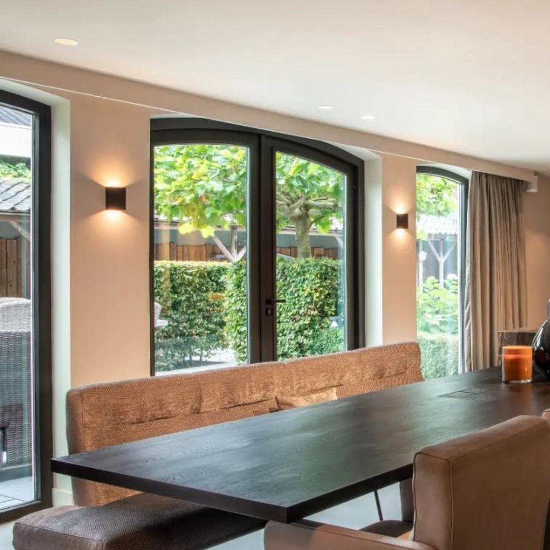 Modern dining room with sleek black table, cushioned bench seating, and large arched windows overlooking a lush garden. Contemporary interior design.