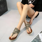 Woman wearing leopard print sandals with white soles, sitting on a gray floor. Fashionable summer footwear, casual style, trendy animal print design.