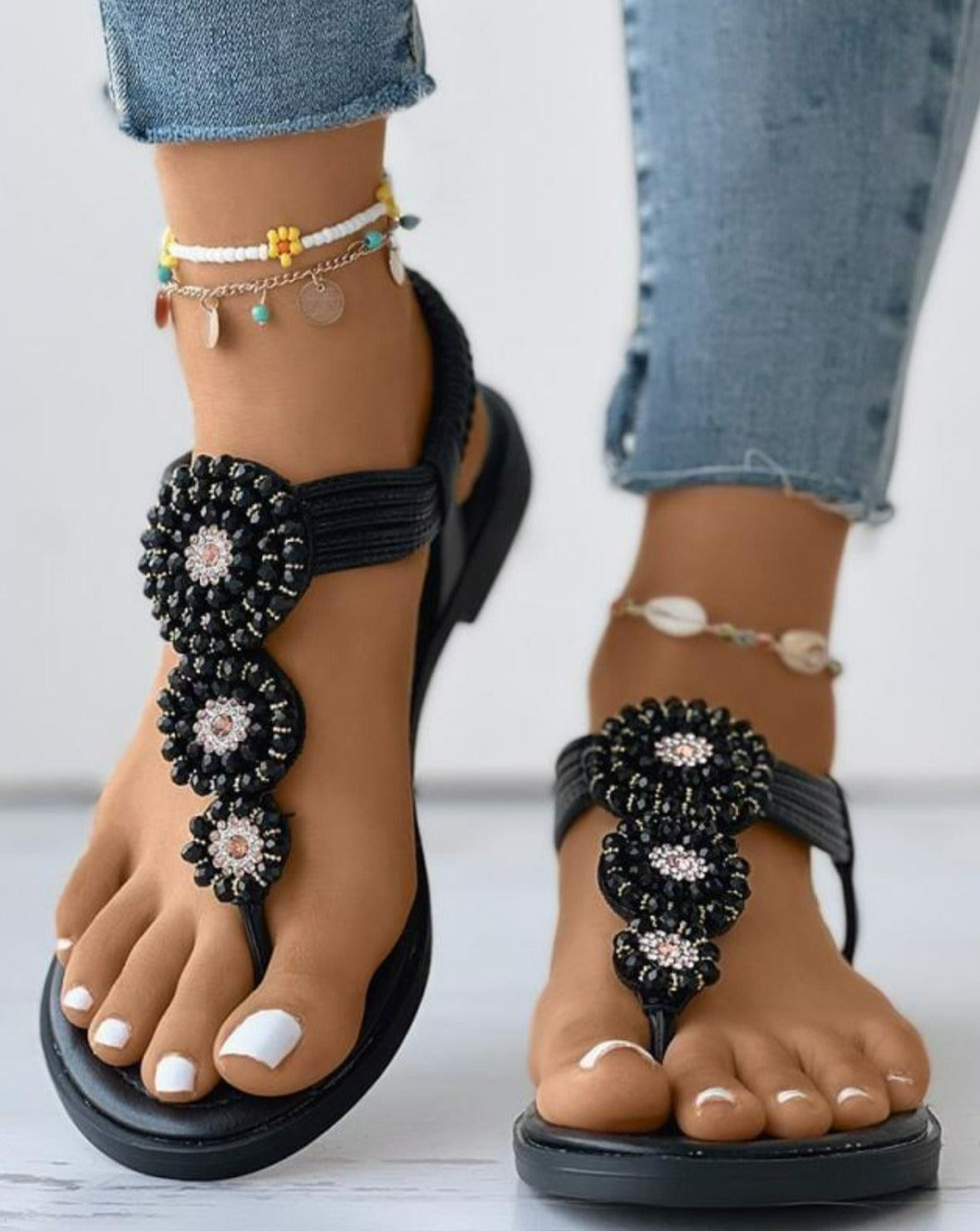 Beaded black sandals with floral design on woman's feet, paired with colorful anklets and jeans. Fashionable summer footwear, boho style.