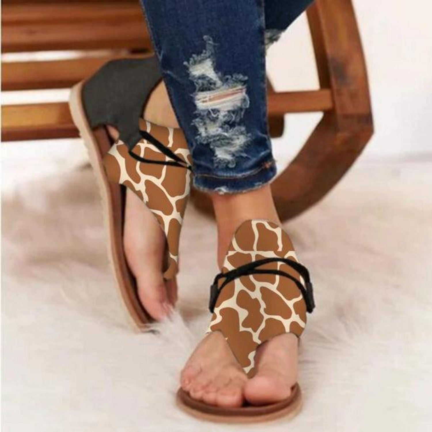 Women's giraffe print sandals with black straps, worn with ripped jeans, on a wooden bench. Stylish summer footwear, casual fashion.