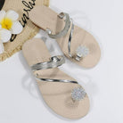 Beige women's sandals with silver rhinestone straps and floral embellishments, displayed on a white background with a straw hat and plumeria flowers.