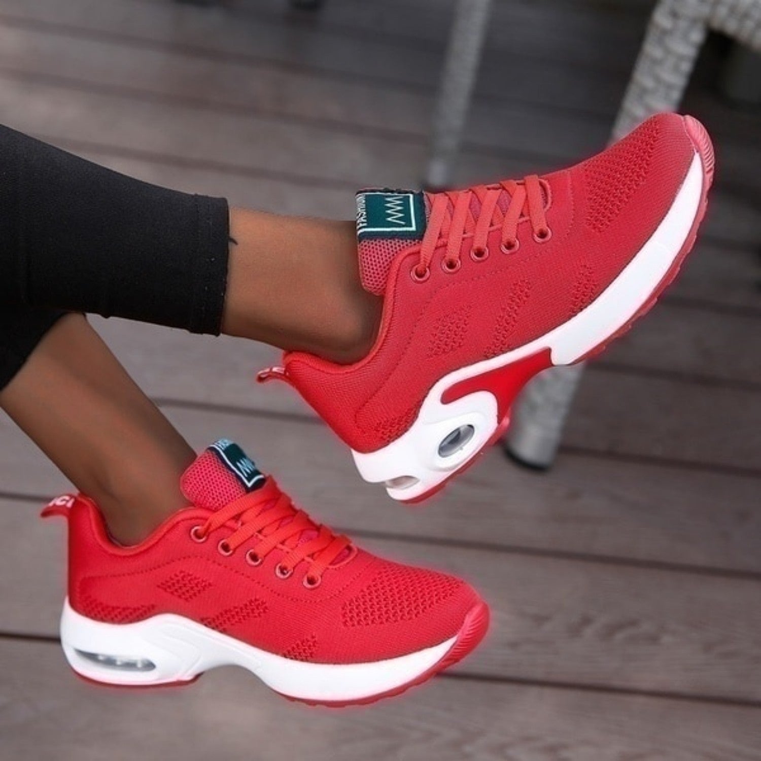 Red athletic sneakers with white soles and air cushioning, worn by a person on a wooden floor. Stylish, comfortable footwear for running or casual wear.