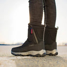 Black winter boots with rugged soles, side zippers, and red-blue pull tabs, worn outdoors on a sunny day. Ideal for cold weather and outdoor activities.