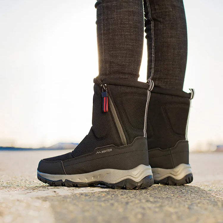 Black winter boots with rugged soles, side zippers, and red-blue pull tabs, worn outdoors on a sunny day. Ideal for cold weather and outdoor activities.