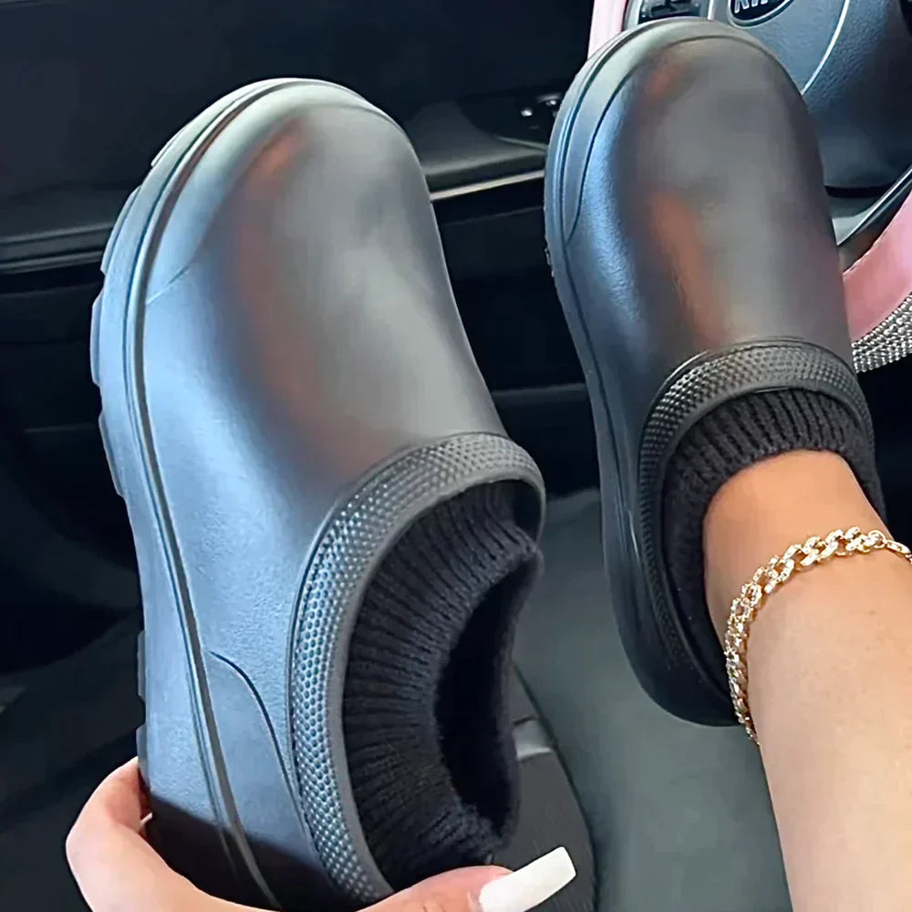 Black slip-on clogs with knit sock lining, held by a person with a gold bracelet, in a car interior. Comfortable, stylish footwear for casual wear.