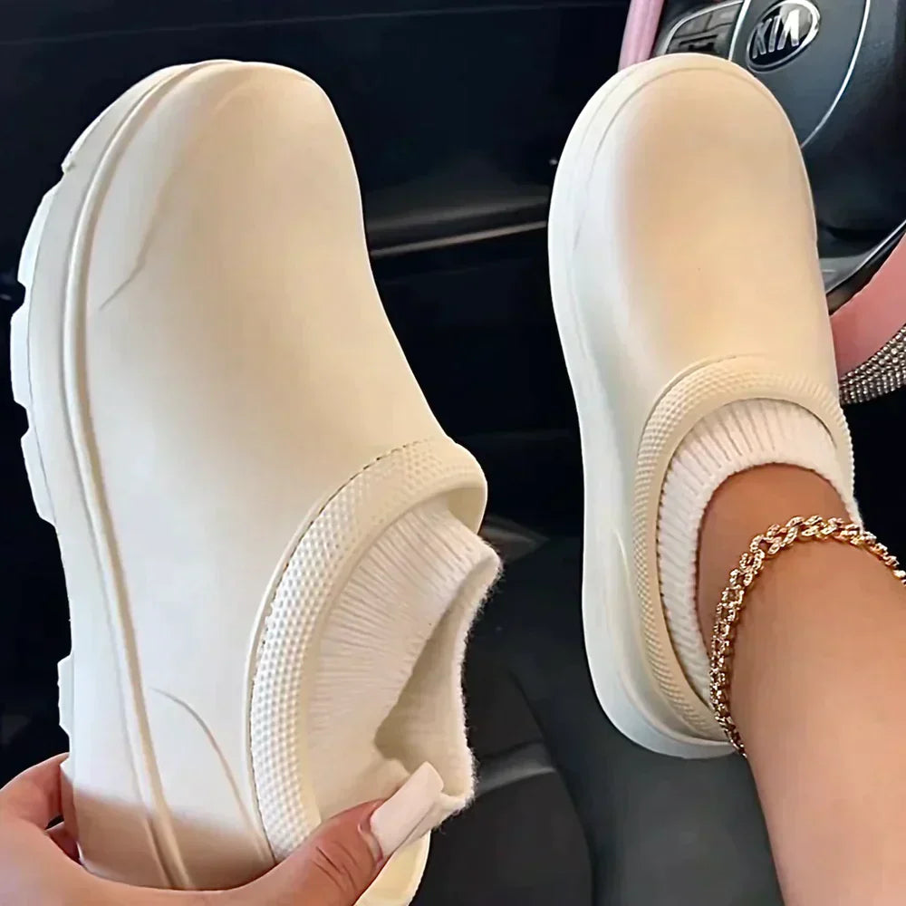 White slip-on clogs with textured soles and ribbed sock-like inserts, worn in a car interior. Fashionable footwear, casual style, comfortable shoes.