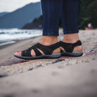 Black strappy sandals on a beach, worn by a person in jeans. Comfortable summer footwear, perfect for casual outdoor activities and beach walks.