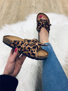 Leopard print sandals with cork sole, held by a person wearing blue jeans and pink nail polish, on a white fluffy rug and wooden floor background.