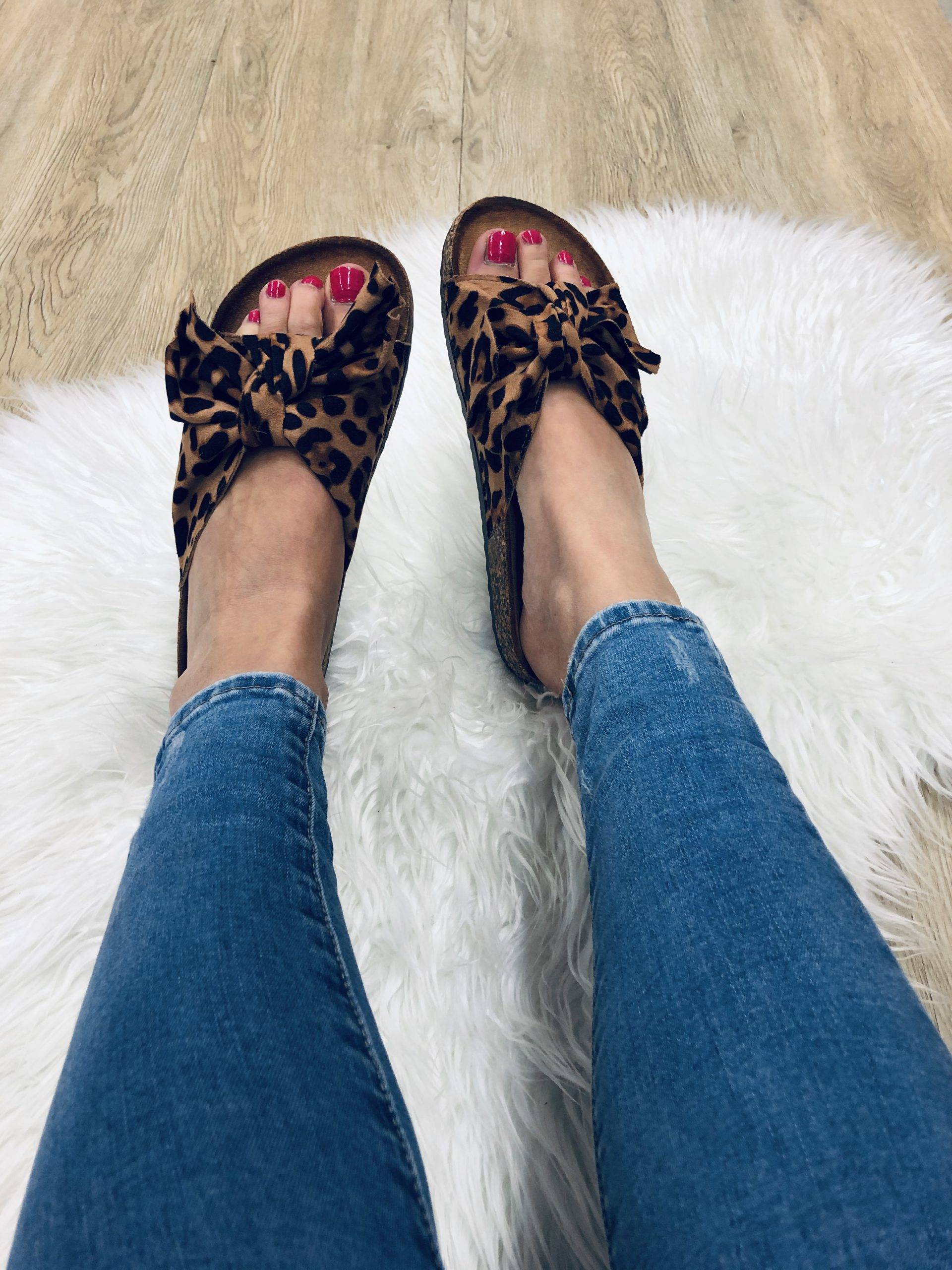 Leopard print bow sandals on woman's feet with red toenails, resting on a white fluffy rug, wearing blue jeans. Fashionable footwear, casual style.