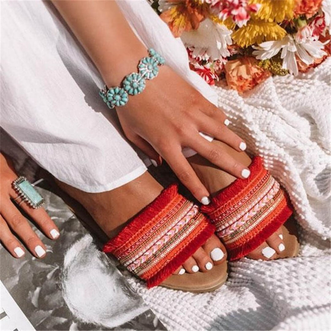 Woman wearing vibrant red sandals with intricate embroidery, turquoise jewelry, and white nail polish, surrounded by colorful flowers on a textured blanket.