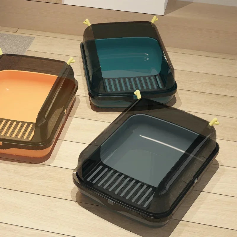 Three enclosed cat litter boxes in orange, teal, and gray with transparent lids on wooden floor.