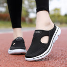 Black slip-on athletic walking shoes with breathable mesh design and white soles, worn outdoors on a red track. Perfect for casual sportswear.