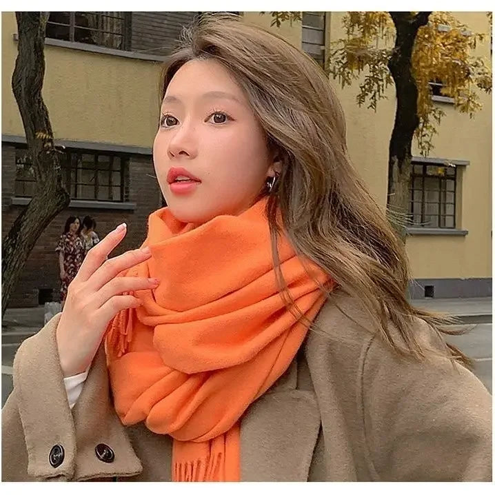 Woman wearing an orange scarf and brown coat, standing outdoors with autumn trees in the background. Fashionable fall outfit, street style.