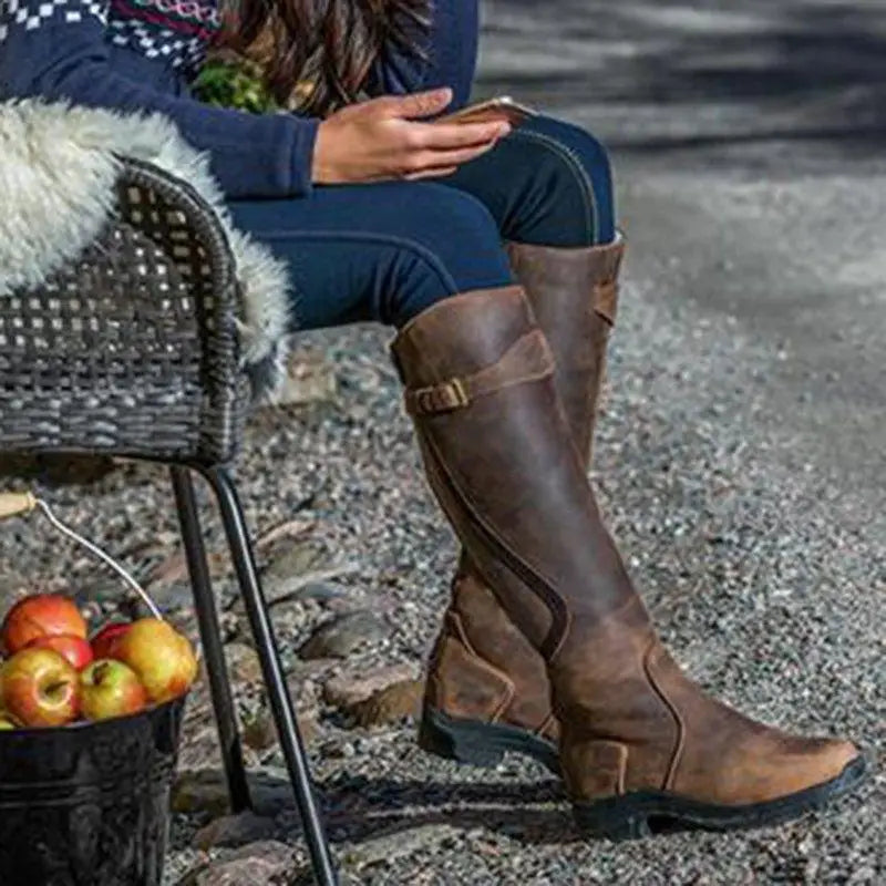 Woman wearing brown leather knee-high riding boots sitting outdoors with a basket of apples. Stylish equestrian footwear, casual fall fashion.