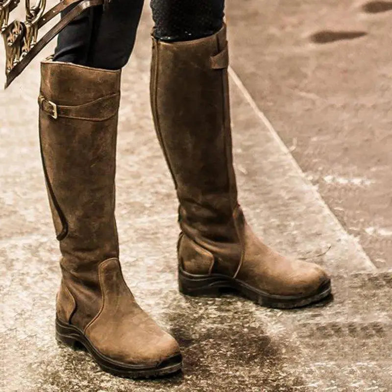 Brown leather knee-high boots with buckle detail on a textured surface, showcasing stylish equestrian fashion. Perfect for fall and winter wear.