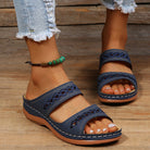 Blue open-toe sandals with decorative stitching on a woman's feet, paired with frayed jeans and a green beaded anklet, on a wooden floor.