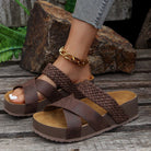 Brown woven platform sandals on a woman's foot, featuring a gold anklet, set against a rustic wooden background with rocks and greenery.