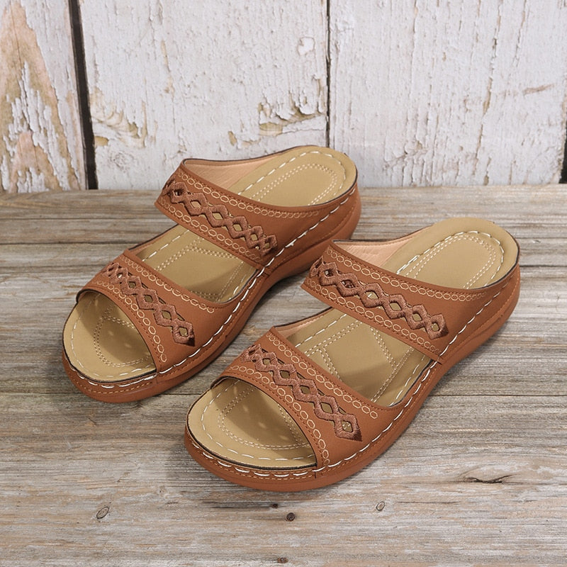 Women's brown leather sandals with decorative stitching, open-toe design, and cushioned sole on rustic wooden background. Summer footwear fashion.