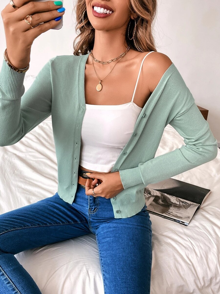 Woman in mint green cardigan, white crop top, and blue jeans sitting on bed, holding phone.