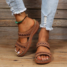 Brown leather sandals with intricate cutout design on woman's feet, paired with ripped jeans and a green beaded anklet, wooden background.