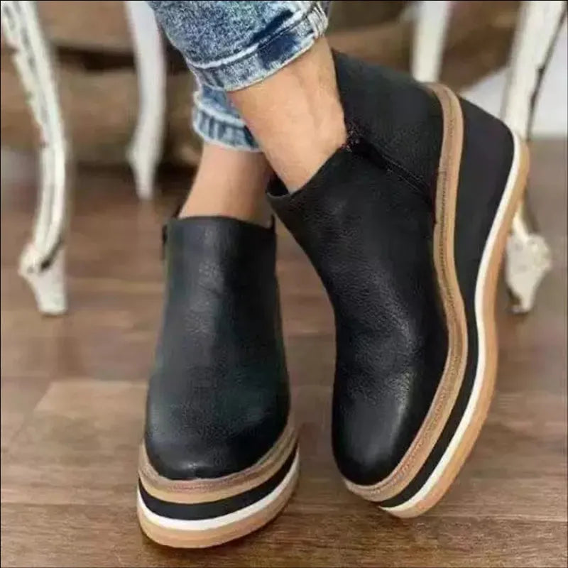 Black leather platform ankle boots with white and brown layered soles, worn with blue jeans on a wooden floor. Fashion footwear, women's shoes.