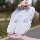 White mesh slip-on sneakers with cushioned soles, held outdoors. Lightweight, breathable design ideal for walking and casual wear.