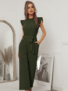 Woman in elegant green jumpsuit with ruffled sleeves and waist tie, standing in stylish room with mirror and decorative elements. Fashionable outfit.