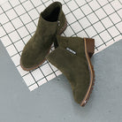 Olive green suede ankle boots with chunky heels on a checkered background, featuring side zippers and stitched detailing. Fashionable women's footwear.