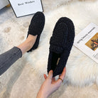 Black fuzzy loafers on a white rug, held by a person wearing jeans. Cozy footwear, stylish design, perfect for casual wear.