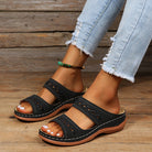 Women's black slip-on sandals with cutout design, worn with distressed light blue jeans. Fashionable summer footwear on wooden floor background.