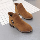 Brown suede ankle boots with side zipper on a gray grid-patterned surface, featuring a chunky heel and stitched detailing. Fashionable women's footwear.