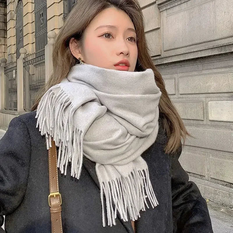 Woman wearing a large gray scarf with fringe, black coat, and brown shoulder bag, standing outdoors against a stone building. Winter fashion style.