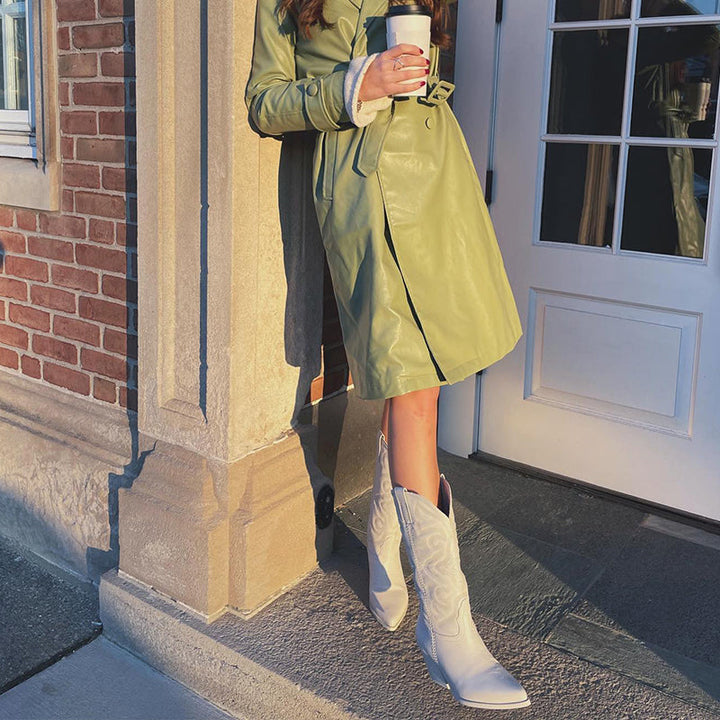 Woman in green trench coat and white cowboy boots holding coffee, standing by a brick wall and door. Fashionable autumn outfit, urban style.