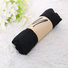 Rolled black t-shirt wrapped in brown paper on white fluffy rug, next to white flowers. Casual clothing, minimalist packaging, cozy home decor.
