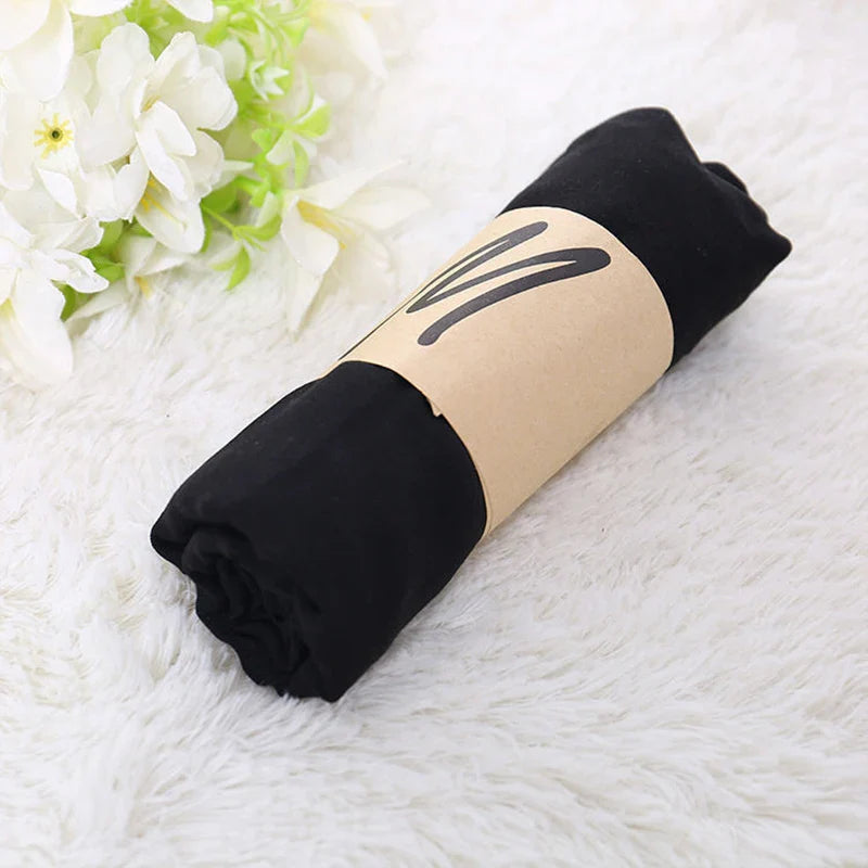 Rolled black t-shirt wrapped in brown paper on white fluffy rug, next to white flowers. Casual clothing, minimalist packaging, cozy home decor.