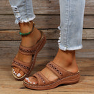 Women's brown leather sandals with cutout design, worn with distressed jeans and a green anklet, on a wooden floor background.