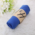 Blue muslin swaddle blanket rolled with a brown paper band, placed on a soft white surface, next to white flowers. Ideal for baby care and nursery use.