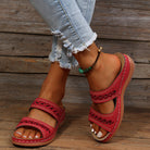 Red leather sandals with cutout design on woman's feet, paired with distressed denim jeans and a beaded anklet, rustic wooden background.