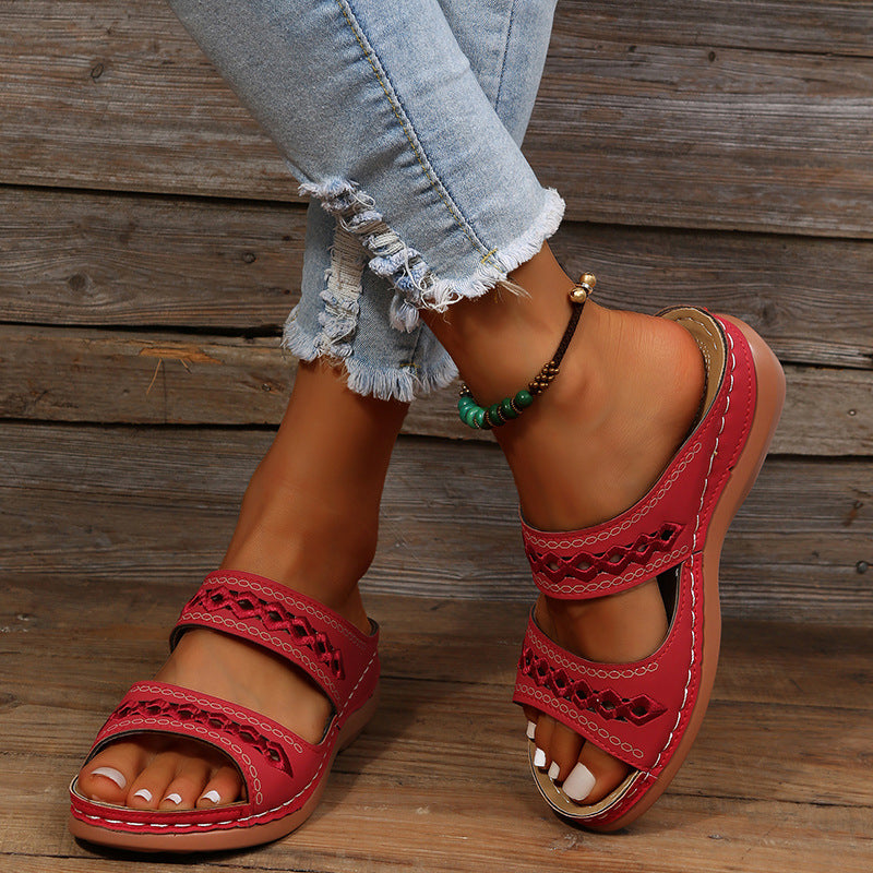 Red leather sandals with cutout design on woman's feet, paired with distressed denim jeans and a beaded anklet, rustic wooden background.