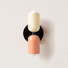 Modern wall sconce with dual-tone cylindrical design, featuring pastel yellow and peach colors, mounted on a black circular base. Contemporary lighting fixture.