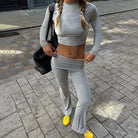 Woman in gray long-sleeve crop top and flared pants, standing on a cobblestone street, wearing yellow sneakers and carrying a black bag.