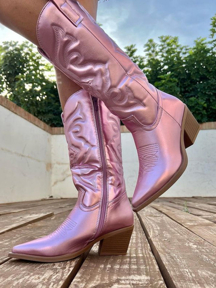 Pink metallic cowboy boots with intricate stitching, mid-calf height, and block heels on a wooden deck. Fashionable western footwear for women.