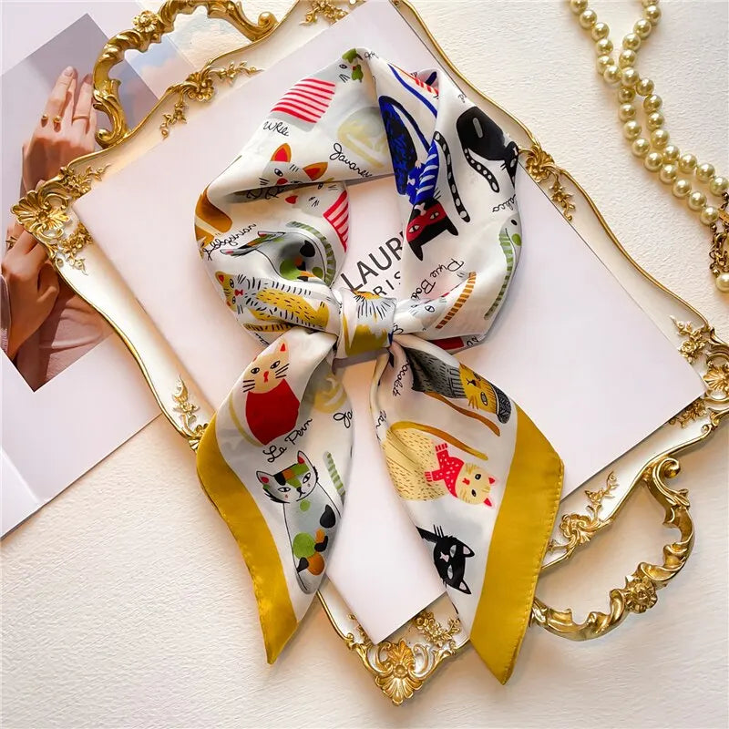 Silk scarf with colorful cat illustrations and yellow border, elegantly displayed on a decorative frame, next to a pearl necklace.
