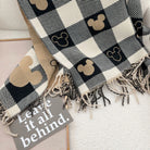Plaid blanket with Mickey Mouse pattern and fringed edges on a white surface, accompanied by a sign reading "Leave it all behind." Cozy home decor.