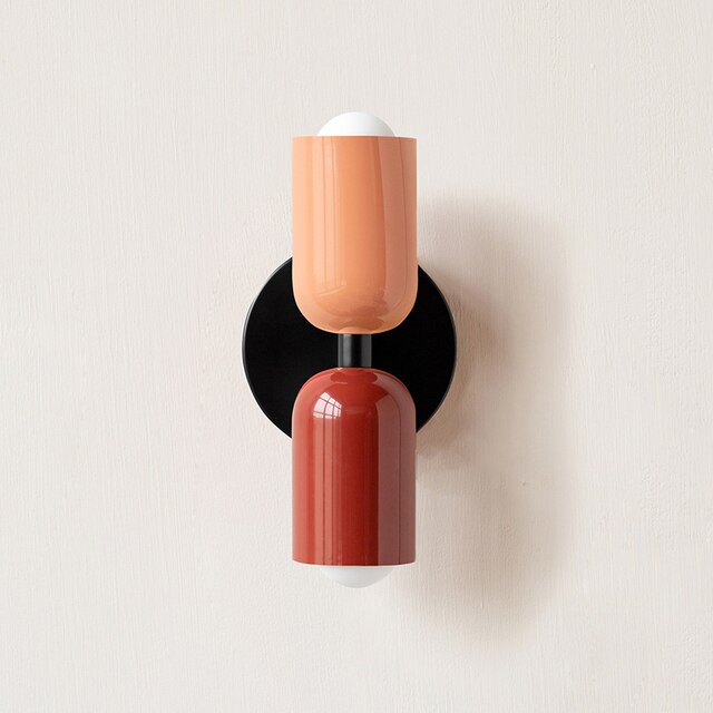 Modern wall sconce with dual-tone cylindrical design in peach and red, mounted on a black circular base. Contemporary lighting fixture for home decor.